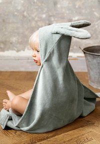 Grå bomullshandduk med kaninmotiv och stora öron, hängande över ett barn som sitter på ett trägolv, som visar mjukt tyg och avslappnad design.
