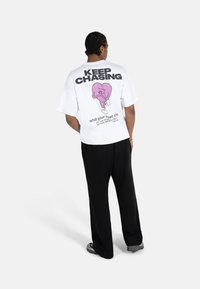 Hvit t-skjorte med et stort grafisk design med et rosa hjerte på baksiden, tekst "KEEP CHASING", kombinert med svarte vide bukser.