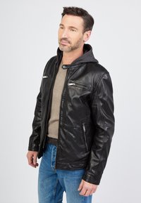 Sort læderjakke med en grå hættetrøje, der har lynlåslommer og en glat struktur. Båret over en beige skjorte og blå jeans.