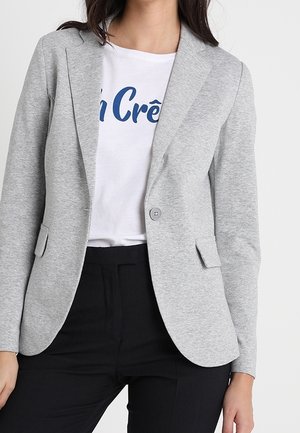 Frau trägt einen hellgrauen Blazer über einem weißen T-Shirt mit blauem Text und schwarze Hose, gezeigt vom Hals bis zu den Hüften.