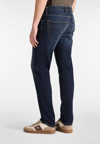 Jeans de mezclilla de color azul oscuro con un corte clásico, que cuentan con cinco bolsillos, hilo en contraste y se combinan con zapatillas beige y marrones.