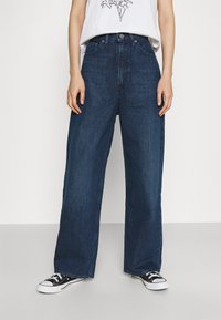 Pessoa vestindo jeans de pernas largas azul escuro, tênis pretos e brancos, e uma blusa branca com uma impressão gráfica parcial contra um fundo liso.