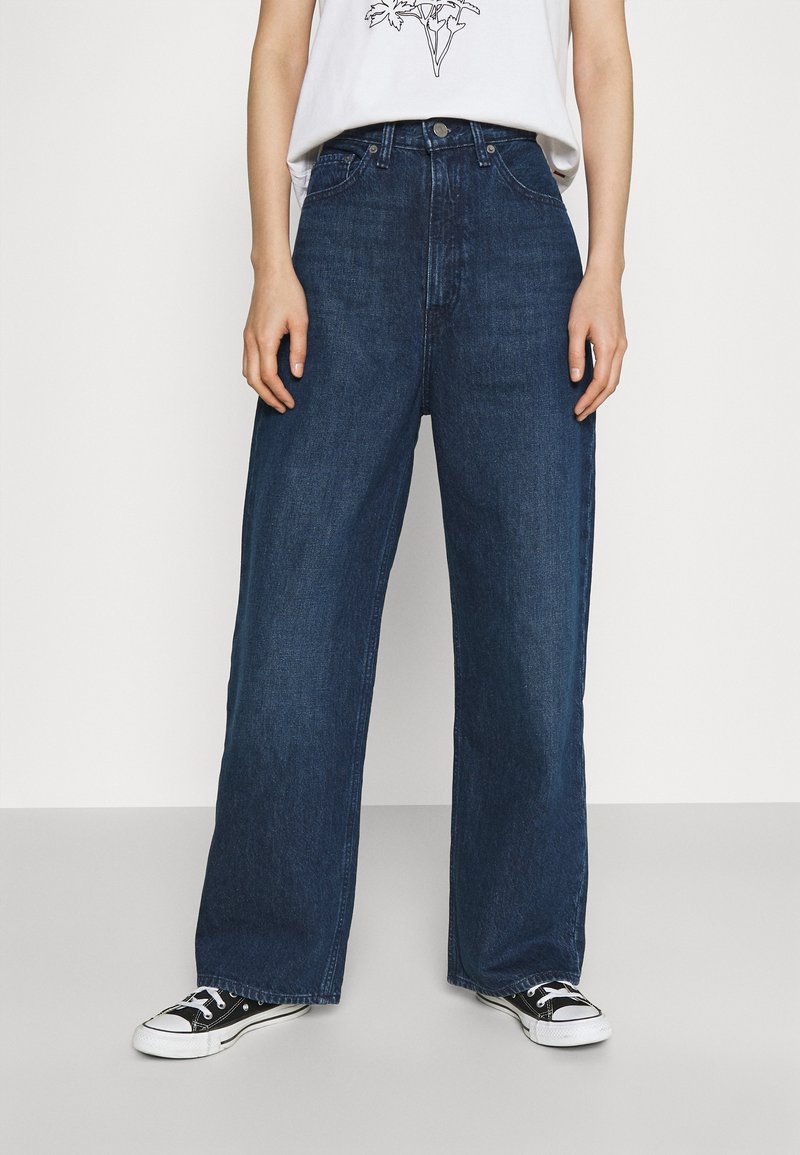 Pessoa vestindo jeans de pernas largas azul escuro, tênis pretos e brancos, e uma blusa branca com uma impressão gráfica parcial contra um fundo liso.