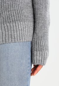 Grauer, gestrickter Pullover mit gerippten Bündchen, der eine sanfte Textur und eine lockere Passform bietet, kombiniert mit hellblauen Jeans.