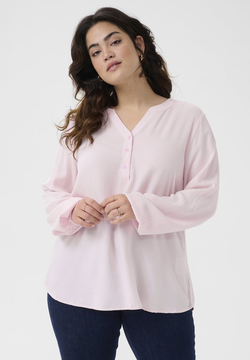 Haut rose clair texturé à manches longues avec un col en V et trois boutons. Tissu doux et coupe décontractée, associé à un jean en denim bleu foncé.