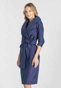 Sukienka w kolorze granatowym i fioletowym, rozkloszowana, z szerokim kołnierzem, rękawami o długości 3/4 oraz wiązaniem w talii, wykonana z gładkiego, błyszczącego materiału.