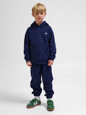 Blond chłopiec stojący, ubrany w granatową bluzę z kapturem i spodnie dresowe z zielonymi tenisówkami, zwrócony przodem na jednolitym jasnoszarym tle.