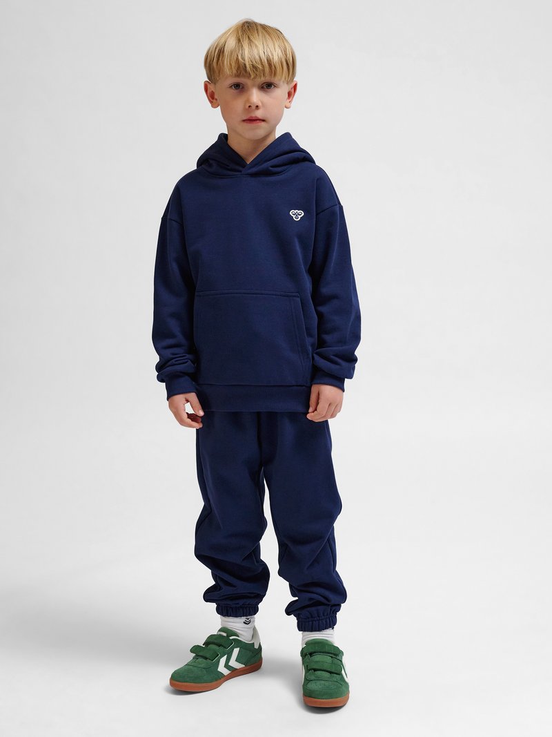 Blonder Junge trägt dunkelblauen Hoodie und Jogginghose mit grünen Sneakers, steht und schaut nach vorne vor schlichtem hellgrauem Hintergrund.