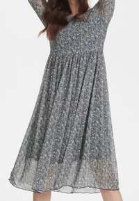 Vestido midi floral con base azul y pequeñas flores amarillas y blancas, mangas largas, cintura fruncida y tela fluida y translúcida.