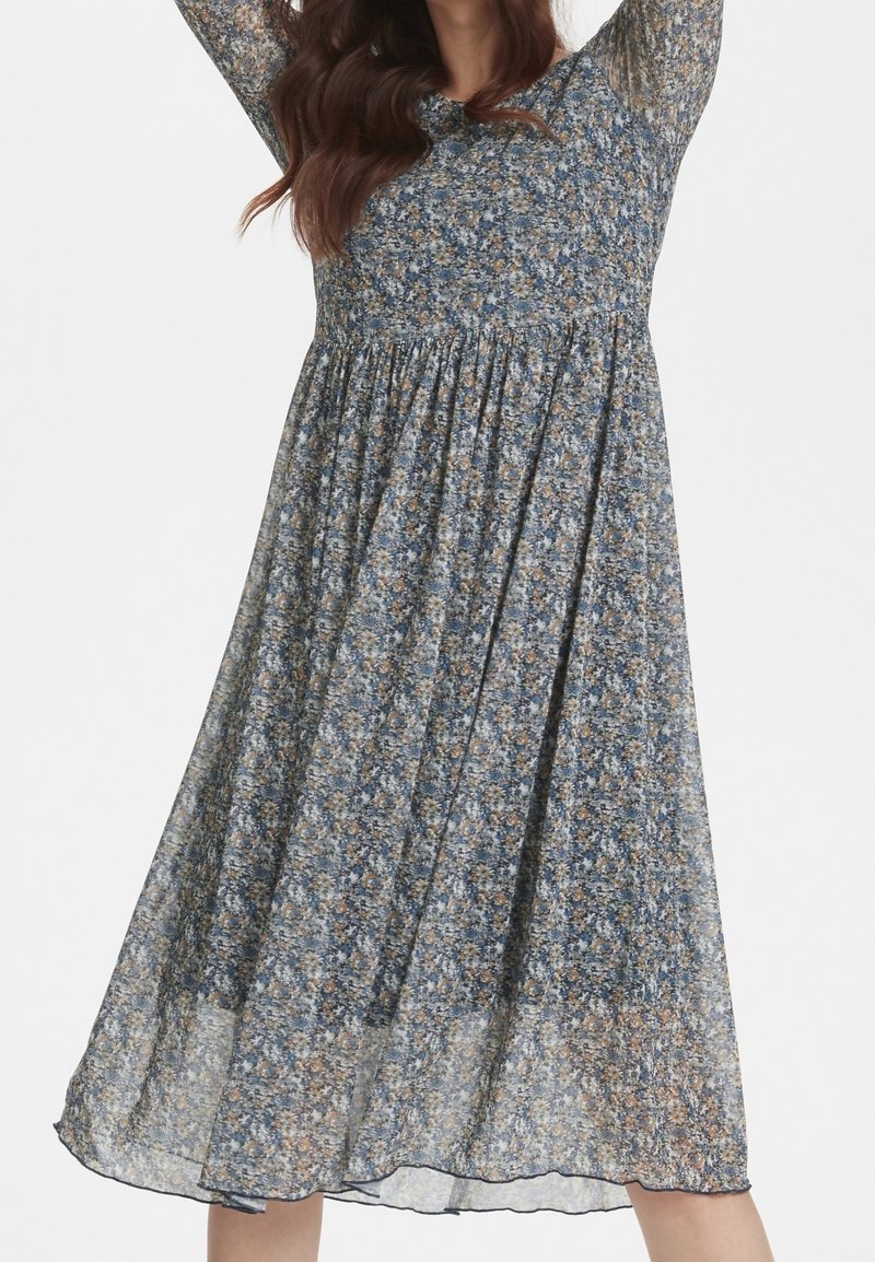 Vestido midi floral con base azul y pequeñas flores amarillas y blancas, mangas largas, cintura fruncida y tela fluida y translúcida.