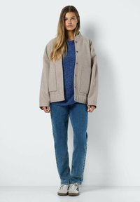 Veste ample beige avec des boutons, poches avant, et un pull en tricot bleu en dessous, associé à un jean bleu clair et des baskets grises.