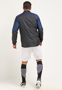 Chaqueta deportiva negra con acentos de rayas azules, combinada con shorts blancos y calcetines negros con rayas horizontales blancas.