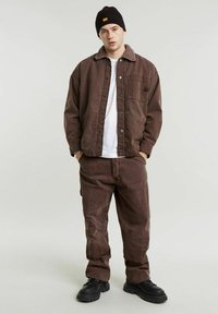 Giacca di tela marrone con un colletto peloso, abbinata a pantaloni coordinati. Indossata sopra una camicia bianca, completata da stivali neri.