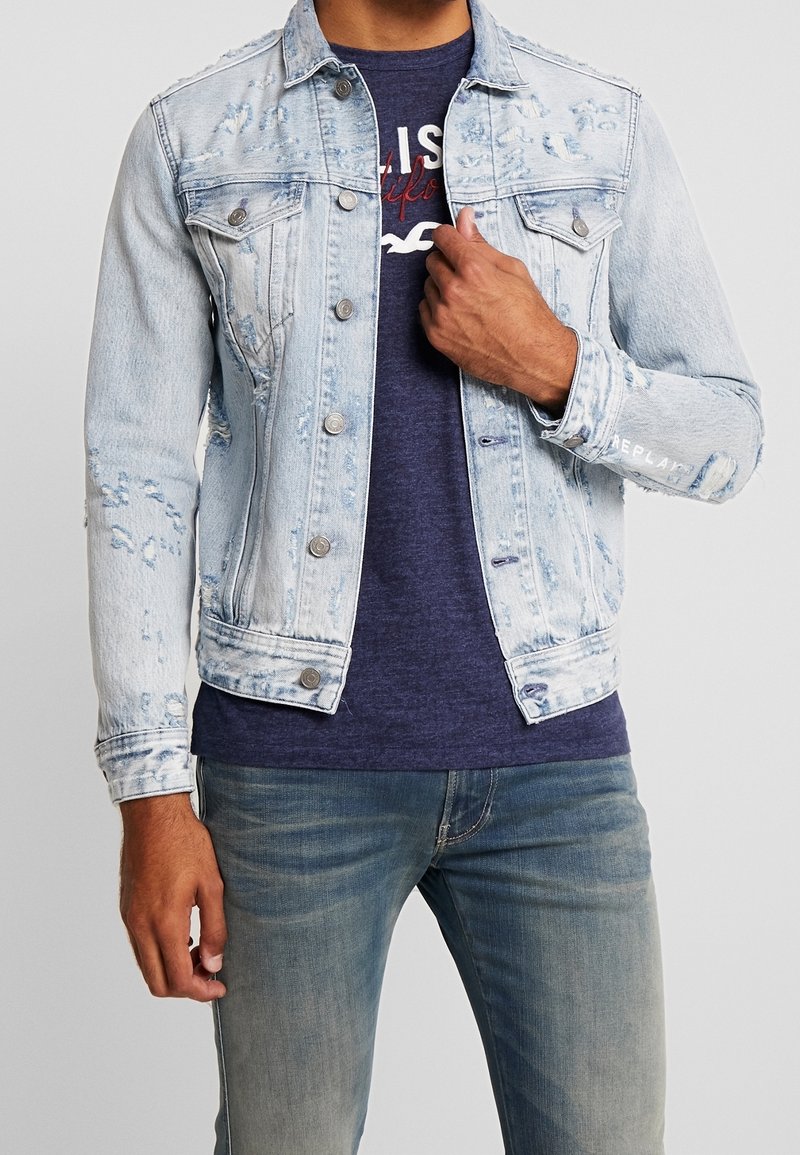 Light blue distressed denim jacket with metal buttons, two chest pockets, and a slightly faded finish, layered over a dark t-shirt.