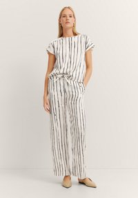 Weißes Outfit mit schwarzen vertikalen Streifen, das kurzärmlig ist und eine gebundene Taille hat, kombiniert mit locker sitzenden Hosen und hellen Schuhen.