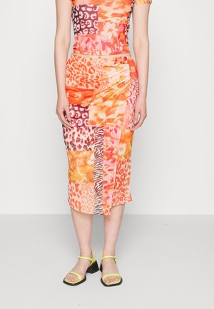 Femme portant une jupe patchwork mi-mollet aux motifs orange, rose et imprimé animal, assortie à des sandales à talons bas avec brides jaunes.