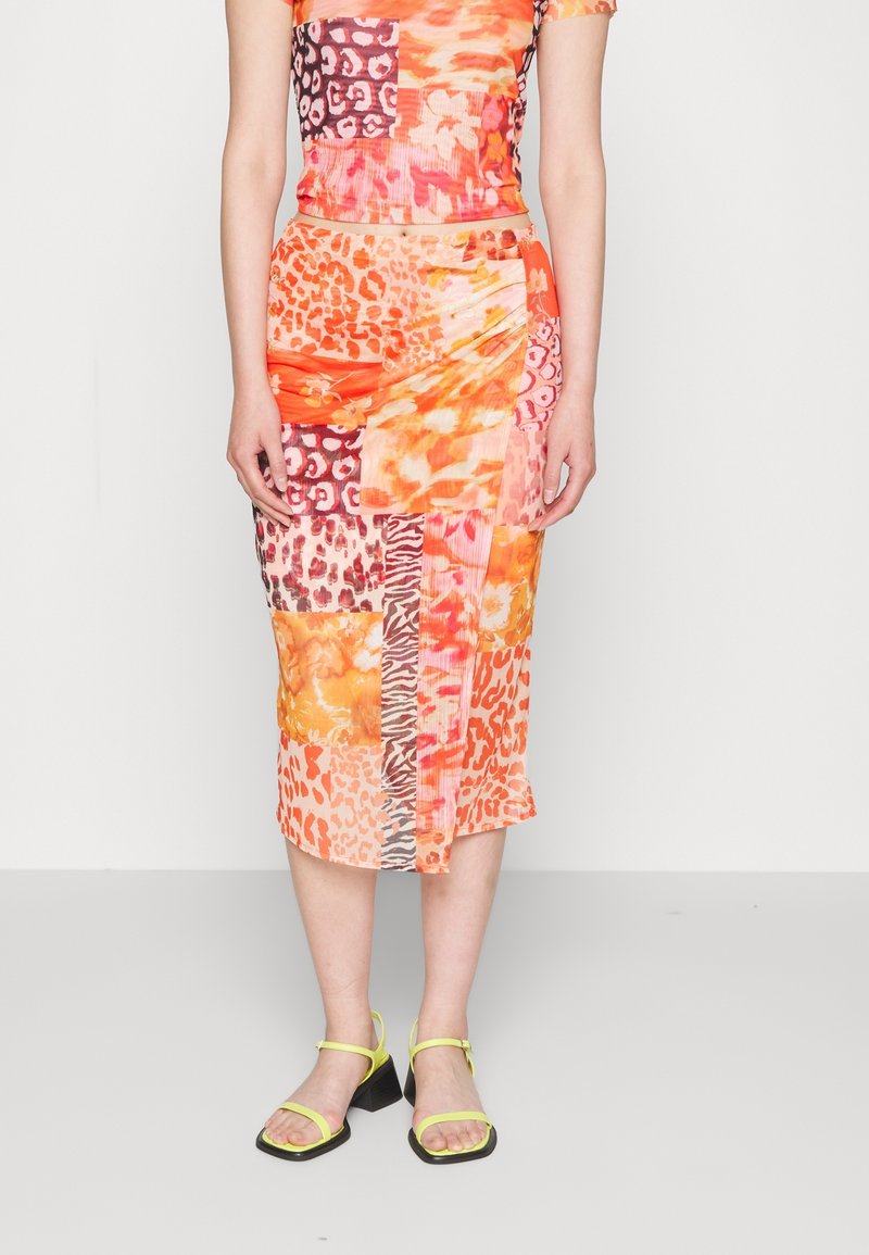 Femme portant une jupe patchwork mi-mollet aux motifs orange, rose et imprimé animal, assortie à des sandales à talons bas avec brides jaunes.