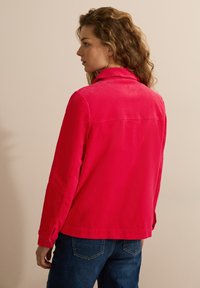 Klarröd kordjacka med krage, långa ärmar och knäppte manschetter; kombinerat med blå jeans, sedd bakifrån.