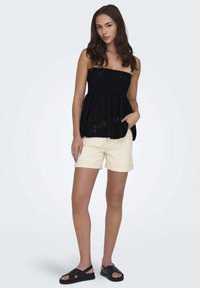 Haut noir sans bretelles en tissu texturé avec des détails en œillets, associé à un short beige clair et des sandales plates noires.