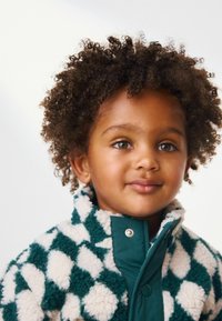 Veste en polaire avec un motif en damier vert et crème, dotée d'un col haut et d'une fermeture à pression. Texture douce, design chaud, adaptée aux enfants.