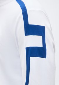 Λευκό ύφασμα με μπλε γεωμετρικό σχέδιο στο πάνω μέρος του βραχίονα που μοιάζει με στυλιζαρισμένο γράμμα Φ ή σχήμα αγκυλής.