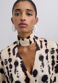 Blusa beige con patrones abstractos en negro y marrón, que presenta un cuello alto y escote en V profundo, combinada con grandes pendientes de aro blancos.