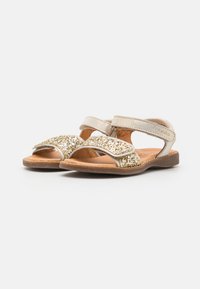 Sandales dorées scintillantes avec une semelle intérieure en cuir marron doux. Elles disposent d'une lanière métallique et d'une fermeture velcro réglable pour un maintien sûr.