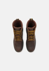 Botas de cuero marrón con cordones amarillos, punta redonda y suela negra robusta. Presentan un collar acolchado y una lengüeta para facilitar su uso.