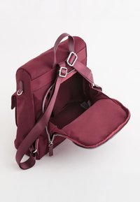 Sac à dos bordeaux de forme rectangulaire, fabriqué en tissu durable. Il dispose de bretelles ajustables, d'une poignée supérieure et d'un compartiment principal zippé.