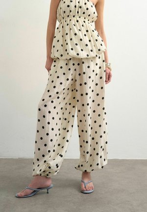 Kvinde iført beige vidde bukser med prikker og elastiske manchetter, kombineret med matchende stropløs top og lyseblå hælede sandaler.