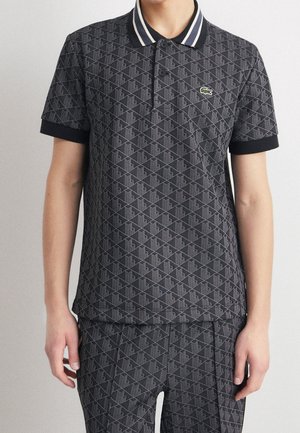 Homme portant un polo noir et un pantalon avec un motif géométrique, un col rayé blanc, et un petit logo crocodile vert sur la poitrine.