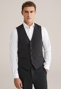Melnā pielāgotā vestē ar teksturētu dizainu, sešām pogām un priekšējo kabatu, uzvalkot virs baltas krekla.