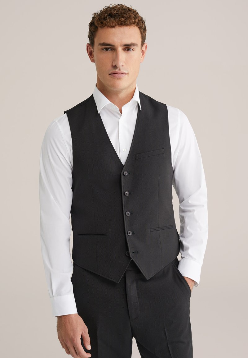 Melnā pielāgotā vestē ar teksturētu dizainu, sešām pogām un priekšējo kabatu, uzvalkot virs baltas krekla.