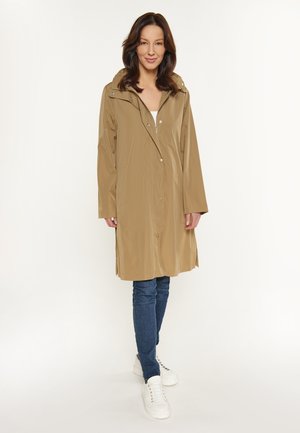 Waterdichte beige regenjas met een capuchon, knopen aan de voorkant en zijzakken, gecombineerd met blauwe spijkerbroeken en witte sneakers. Soepele, lichte textuur.