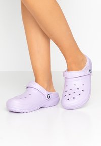 Zoccoli Crocs di colore viola chiaro con cinturino sul retro, fori di ventilazione e suola testurizzata. Realizzati in materiale stampato con una finitura liscia.