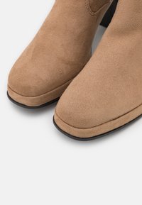 Botine din suede bej, cu vârful rotund, textură netedă și talpă neagră. Au un cusut minim și fără elemente de prindere vizibile.