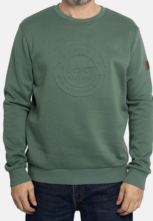 Homme portant un sweat-shirt vert avec un logo circulaire en relief et un jean bleu foncé, les mains détendues le long du corps sur un fond uni.