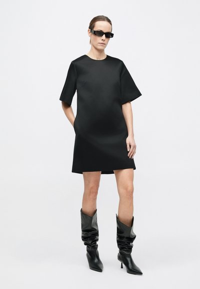 Sort kortærmet kjole med afslappet silhuet og glat tekstur, kombineret med knælange, slaskede sorte støvler og kantede sorte solbriller.