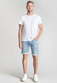 Camiseta de algodón blanca combinada con pantalones cortos de mezclilla claros, con los puños doblados, y zapatillas blancas. El modelo está de pie frente a un fondo neutro.
