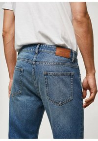 Jean en denim bleu clair avec un design classique à cinq poches, comprenant une étiquette en cuir marron sur la ceinture arrière, et un tissu texturé.