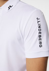 Valkoinen poolopaita, joka on valmistettu sileästä materiaalista. Rinnassa on musta logo ja hihaan on kirjailtu nimi "J.LINDEBERG". Muotoiltu malli.