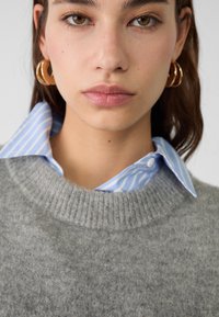 Maglione grigio in lana con scollatura a costine indossato sopra una camicia colletto a righe azzurre. Orecchini a cerchio d'oro visibili.