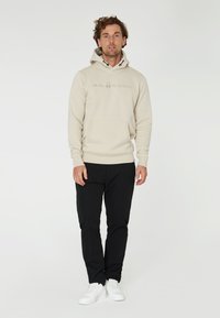 Sudadera beige con cordones, bolsillo frontal y logo bordado; combinada con pantalones negros y zapatillas blancas, sobre un fondo sencillo.