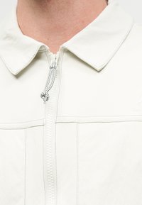 Κοντινό ενός λευκού μπουφάν με γιακά και κεντρικό φερμουάρ με μαύρο-άσπρο δεμένο κορδόνι.