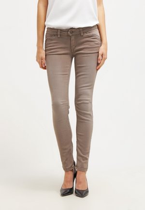 Frau trägt taupefarbene Skinny Jeans und schwarze spitze High Heels, steht vor einem schlichten hellen Hintergrund.