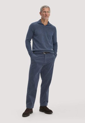 Mann mit grauen Haaren und Brille steht mit den Händen in den Taschen, trägt einen blauen Pullover, blaue Hose, schwarzen Gürtel und dunkelbraune Schuhe.