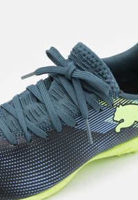 Scarpa sportiva con tomaia in nylon blu scuro texture, strisce blu degradè, lacci elastici e un logo giallo brillante sul lato.
