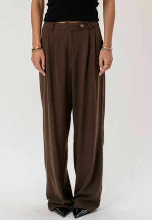 Kobieta nosząca brązowe spodnie z wysokim stanem i szerokimi nogawkami z zapięciem na guziki, w zestawie z czarnymi spiczastymi butami, bransoletkami i pierścionkami.