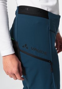 Blauwe sportbroek met zwarte tailleband en ritszak, met het VAUDE-logo. Gemaakt van een gestructureerd, rekbaar materiaal.