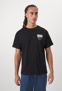 Tricou negru din bumbac cu mâneci scurte, având un grafic alb imprimat cu "1894" și "House of Superfine" pe piept. Design simplu.